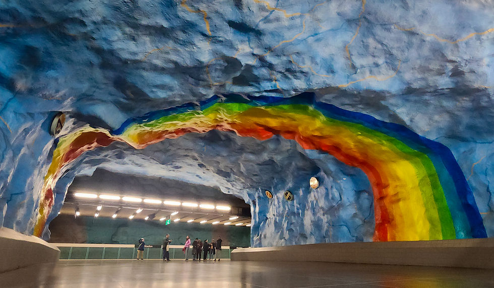 Colorful rainbow mural on a blue cave-like metro station ceiling. People stand below, admiring the art. Bright lights create a vibrant scene.