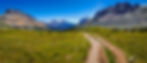 Helen Lake trail leading through green meadow with rocky mountains under a clear blue sky. Snow patches on peaks, serene and picturesque landscape.