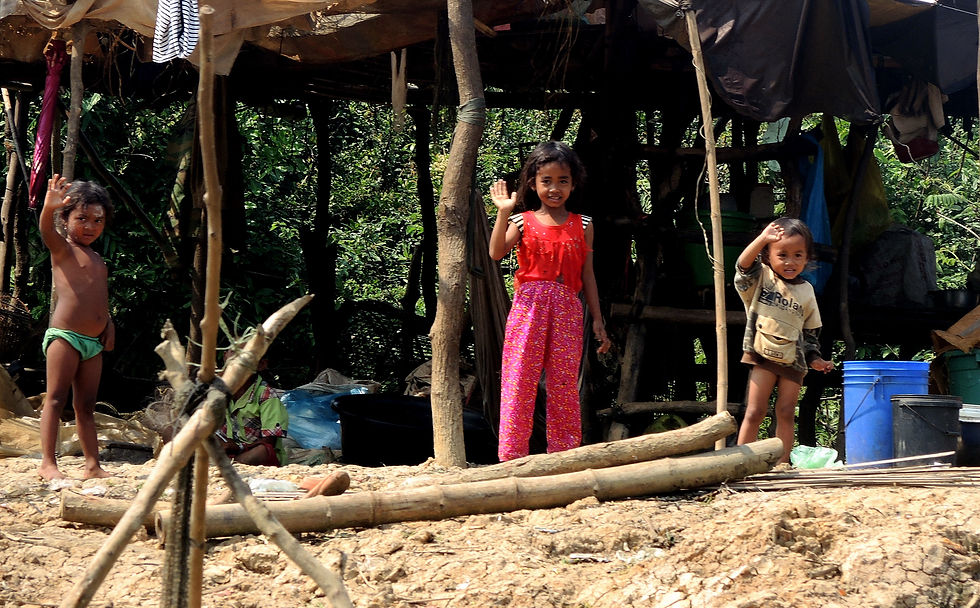 Three children wave outside a makeshift shelter in a forest. The child in red smiles, with trees and scattered items in the background.
