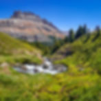 Two people hiking on a grassy hill with a flowing stream. Rugged mountain and pine trees in background under a clear blue sky.
