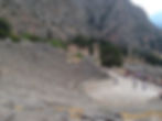 Ancient Delphi amphitheater with stone seats, surrounded by trees and mountains. Tourists explore the circular stage under cloudy skies.