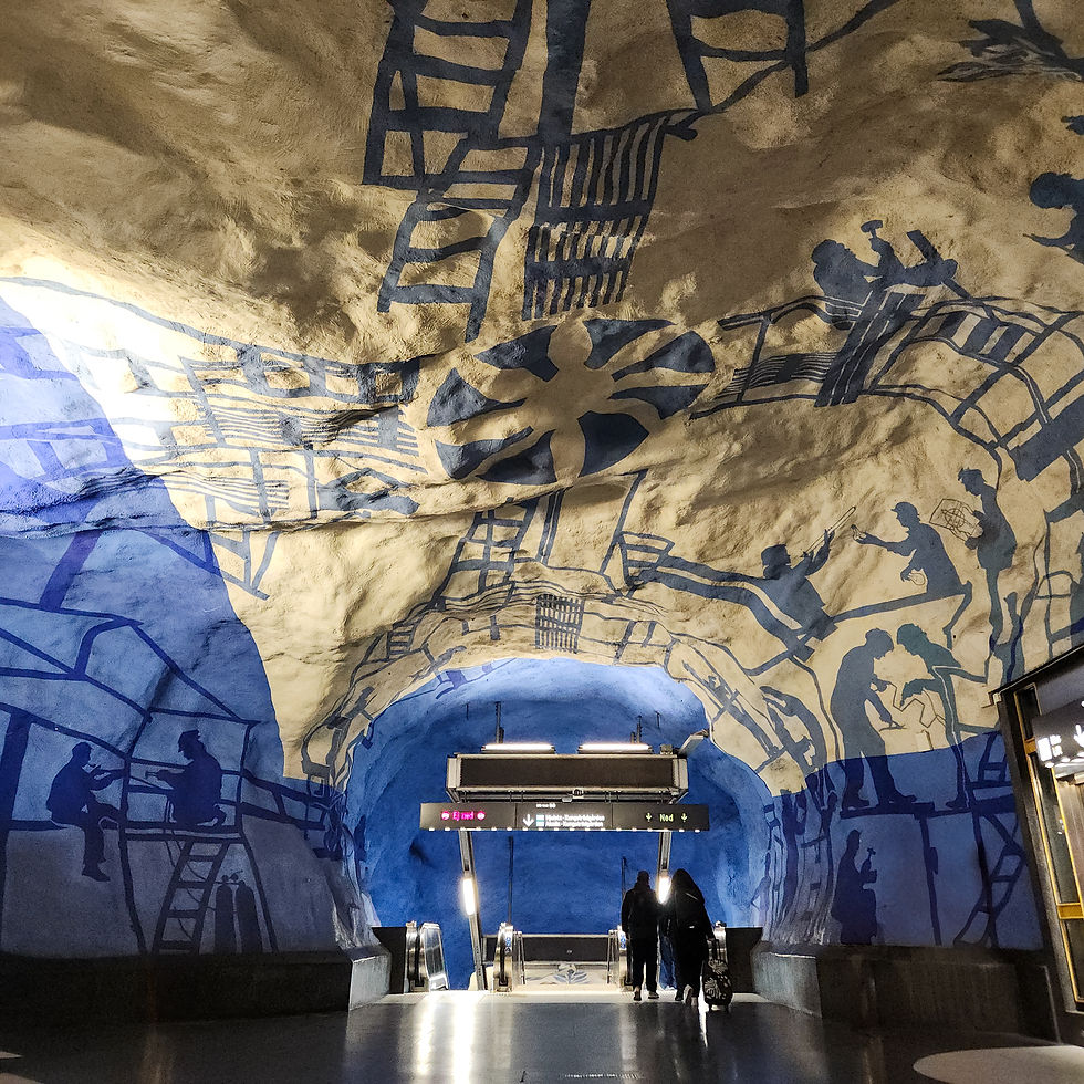 People descend an escalator in a subway with blue and white cave-like murals. Overhead lights illuminate the vibrant artwork.