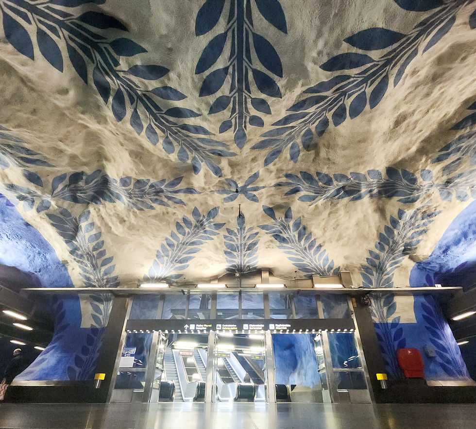 Underground station with blue leaf murals on walls and ceiling. Three escalators lead upwards. Bright lighting creates a serene mood.
