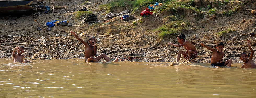 Boys joyfully splash and wave in a muddy river near a grassy, debris-strewn bank under a sunny sky, conveying a playful mood.