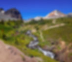 Hikers walk along a lush green trail beside a cascading stream in a mountainous landscape under a clear blue sky. Snow-capped peaks in view.