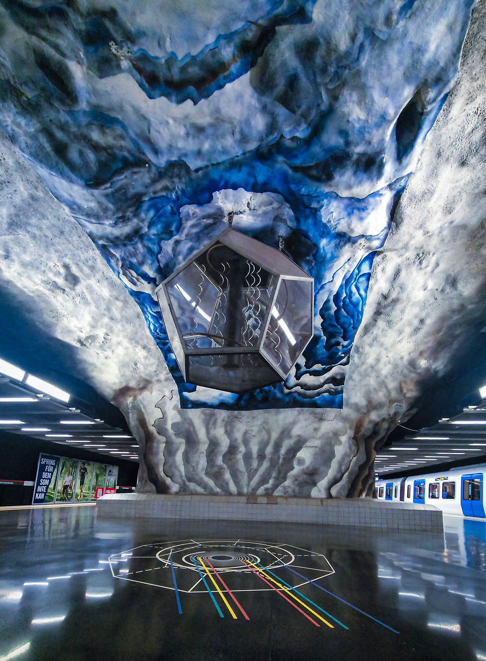 Blue and gray cave-like subway station with hexagonal fixture, colorful floor lines, and a train on the right. Futuristic and artistic.