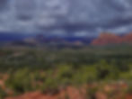 Dramatic, cloudy sky over red rock mountains and lush green trees in a vast landscape, conveying a moody and serene atmosphere.