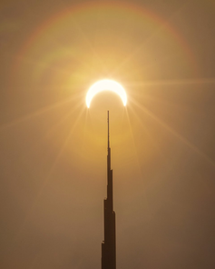 Fotografia simétrica de um eclipse solar anular ("Anel de Fogo") perfeitamente alinhado com o pináculo do Burj Khalifa em Dubai. A imagem exibe tons dourados e ocre, simbolizando a união entre a engenharia humana de ultra-luxo e eventos astronômicos raros.