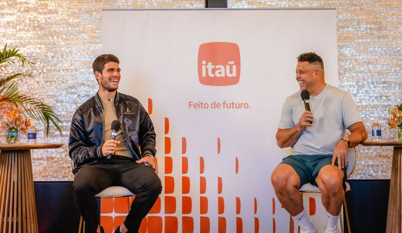 Fotografia de um evento em um ambiente elegante e iluminado. O tenista Carlos Alcaraz (à esquerda) e o ex-jogador de futebol Ronaldo Fenômeno (à direita) estão sentados em banquetas, segurando microfones e rindo descontraidamente. Entre eles, um backdrop branco com detalhes em laranja exibe o logo do Itaú e o slogan "Feito de futuro".