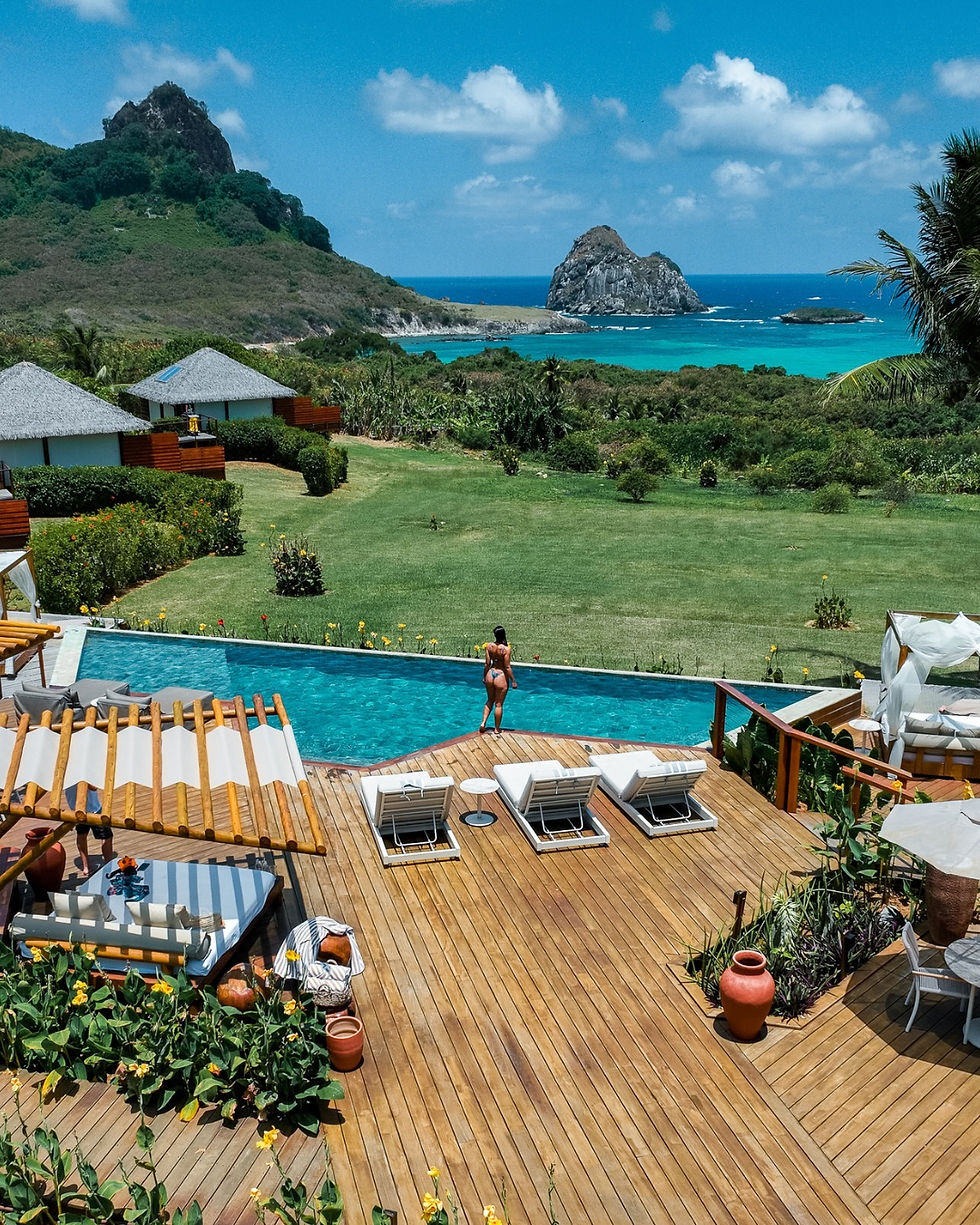 Vista panorâmica de um resort com deck de madeira, piscina azul e gramado verde, tendo o Morro do Pico em Fernando de Noronha ao fundo.