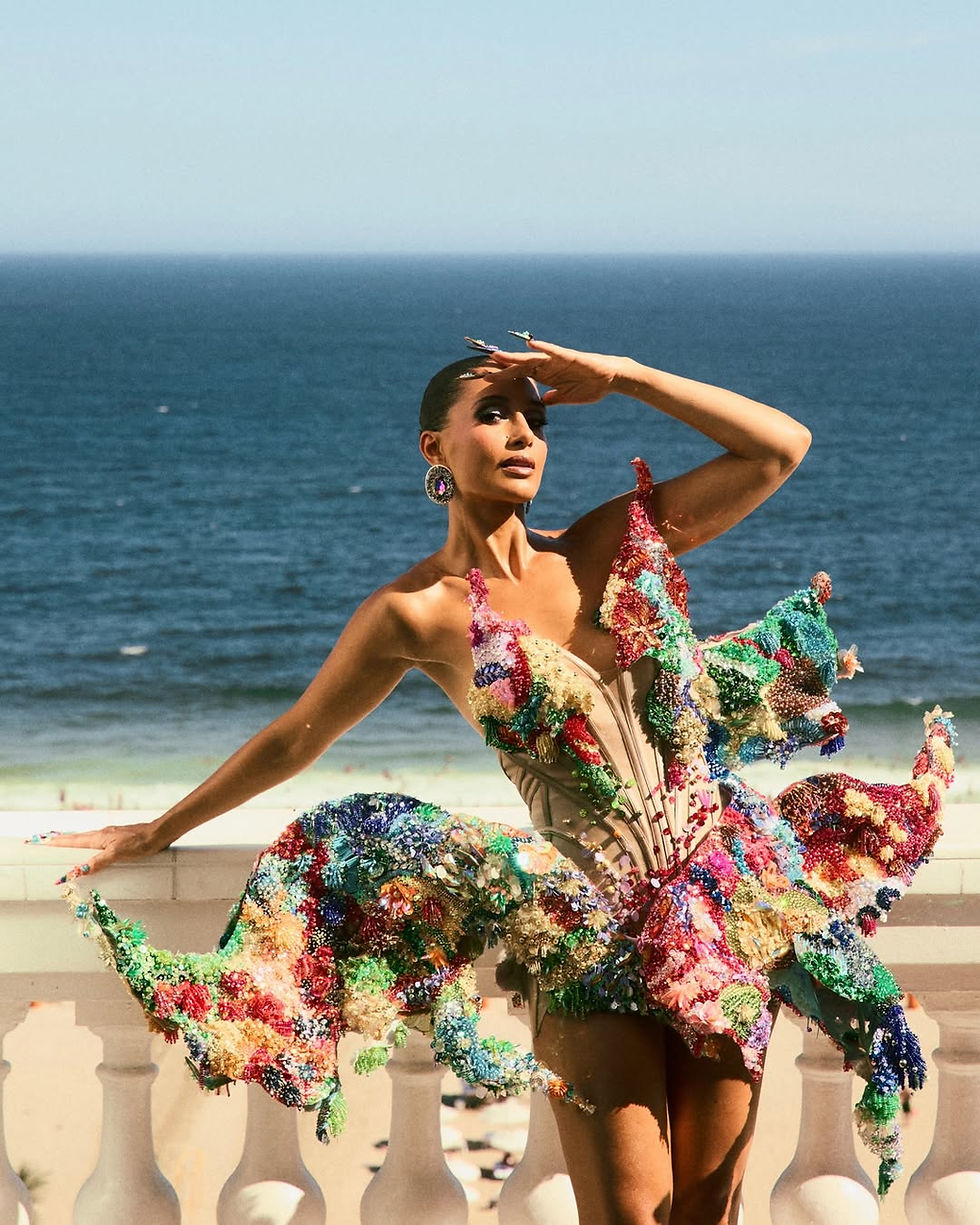 A atriz Camila Pitanga banhada pela luz do sol, posando majestosamente na icônica varanda de balaustrada branca do Copacabana Palace, com a areia da praia e o oceano azul desfocados ao fundo. Ela veste uma criação de alta-costura vanguardista e escultural, com um corset estruturado e inteiramente bordado com milhares de pedrarias, lantejoulas e cristais multicoloridos. A imagem ilustra o contraste entre o isolamento exclusivo e a blindagem do Copacabana Palace no Carnaval e a imensidão da praia.