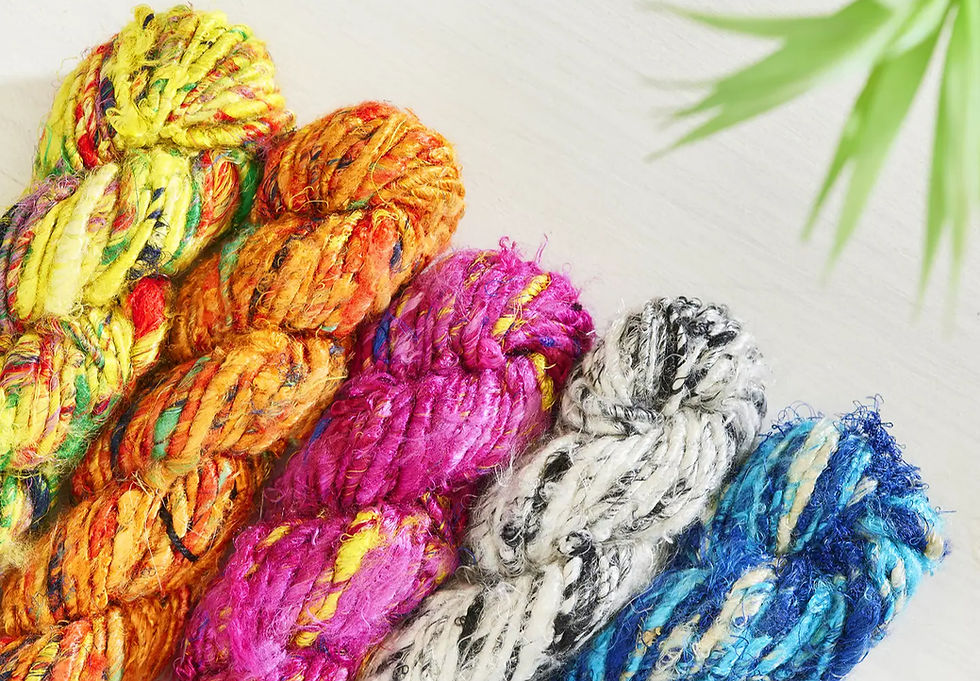 Colorful skeins of yarn in yellow, orange, pink, gray, and blue on a light surface. Green plant leaves in the top right. Vibrant and textured.