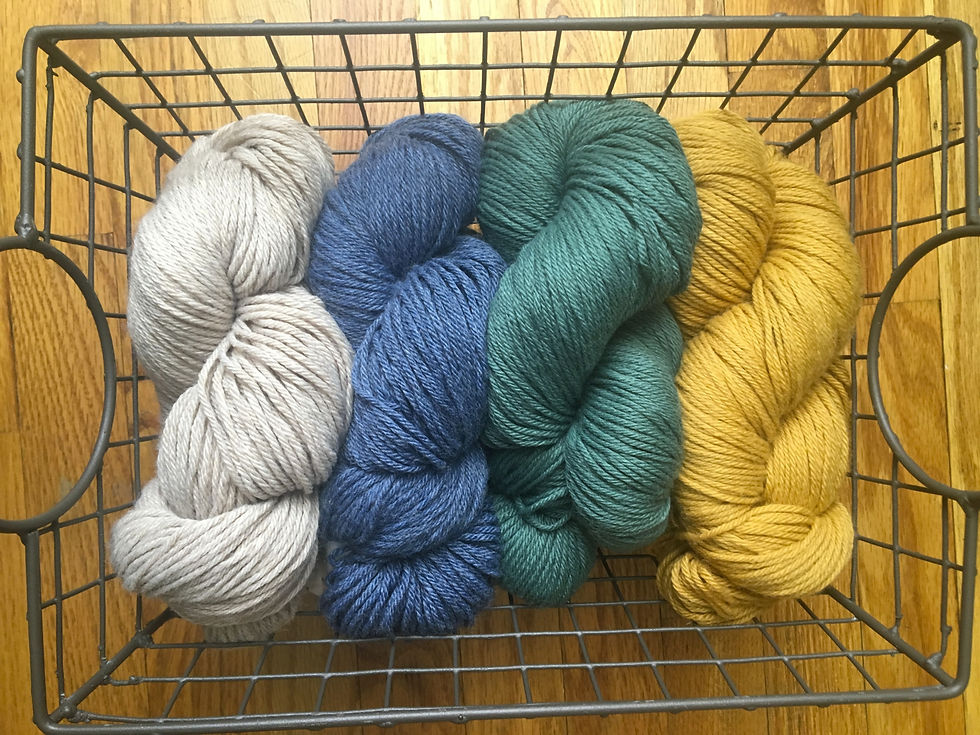 Four skeins of yarn in beige, blue, green, and mustard are arranged in a metal basket on a wooden floor, creating a cozy, creative vibe.