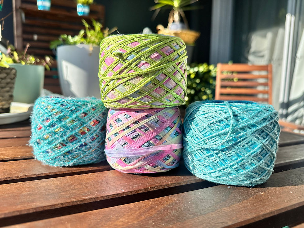 Four vibrant yarns, blue and multicoloured, on wooden table in garden setting. Sunlit plants and a chair in the background.