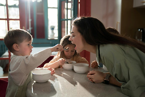 Little boy feeding his mom at the kitchen counter while siblings enjoy breakfast together.