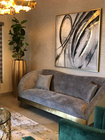 Living visto de perfil. Sillón aterciopelado gris con detalles en dorado. Macetero dorado gigante. De fondo Cuadro de arte abstracto negro con dorado y blanco. 