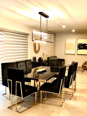 Comedor negro con dorado, alrededor 6 sillas cuadradas negras con aplicaciones doradas. Sobre comedor colgando un candil rectangular linear negro y dorado. 