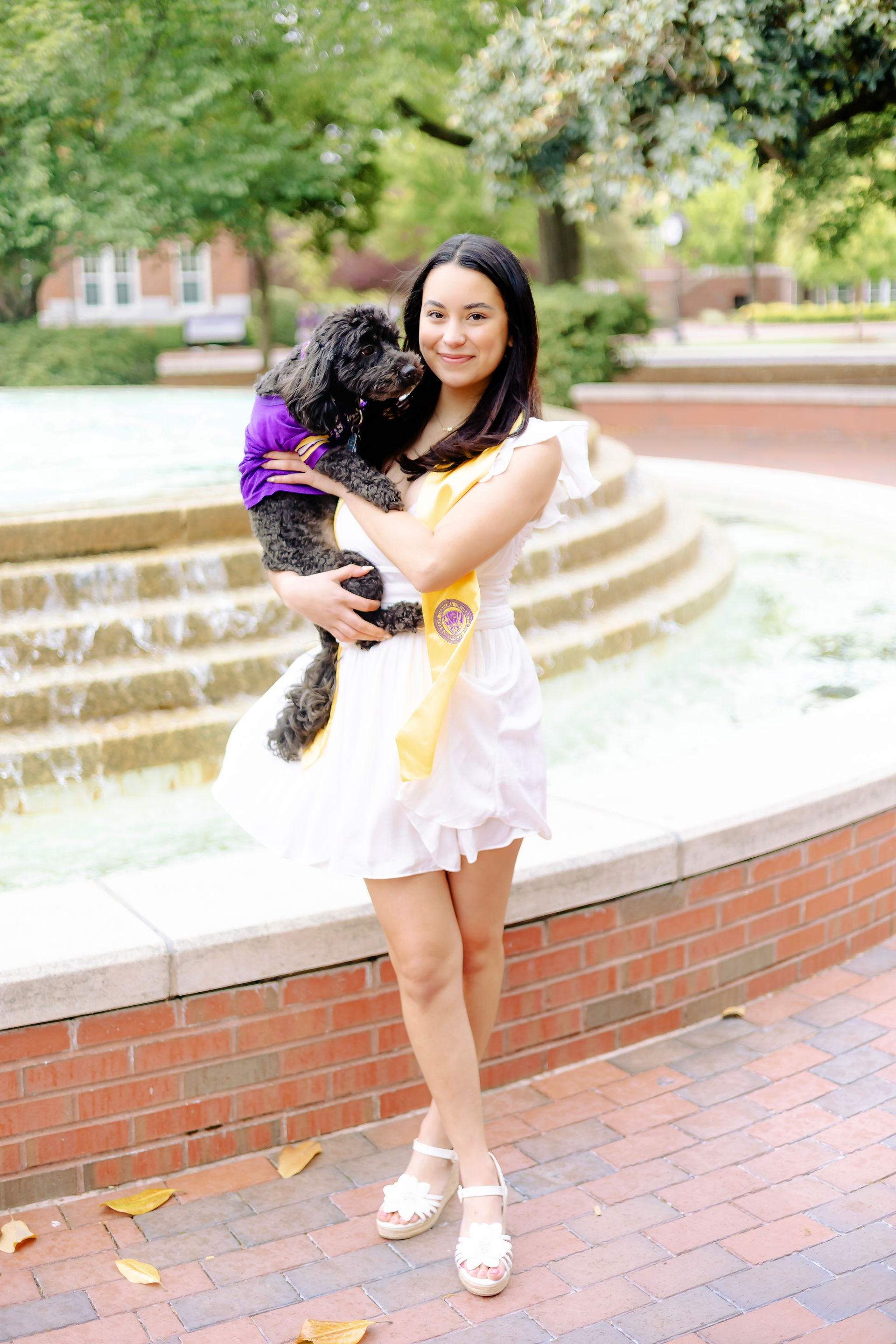ECU senior holding small black dog near East Carolina University fountain during graduation photos.