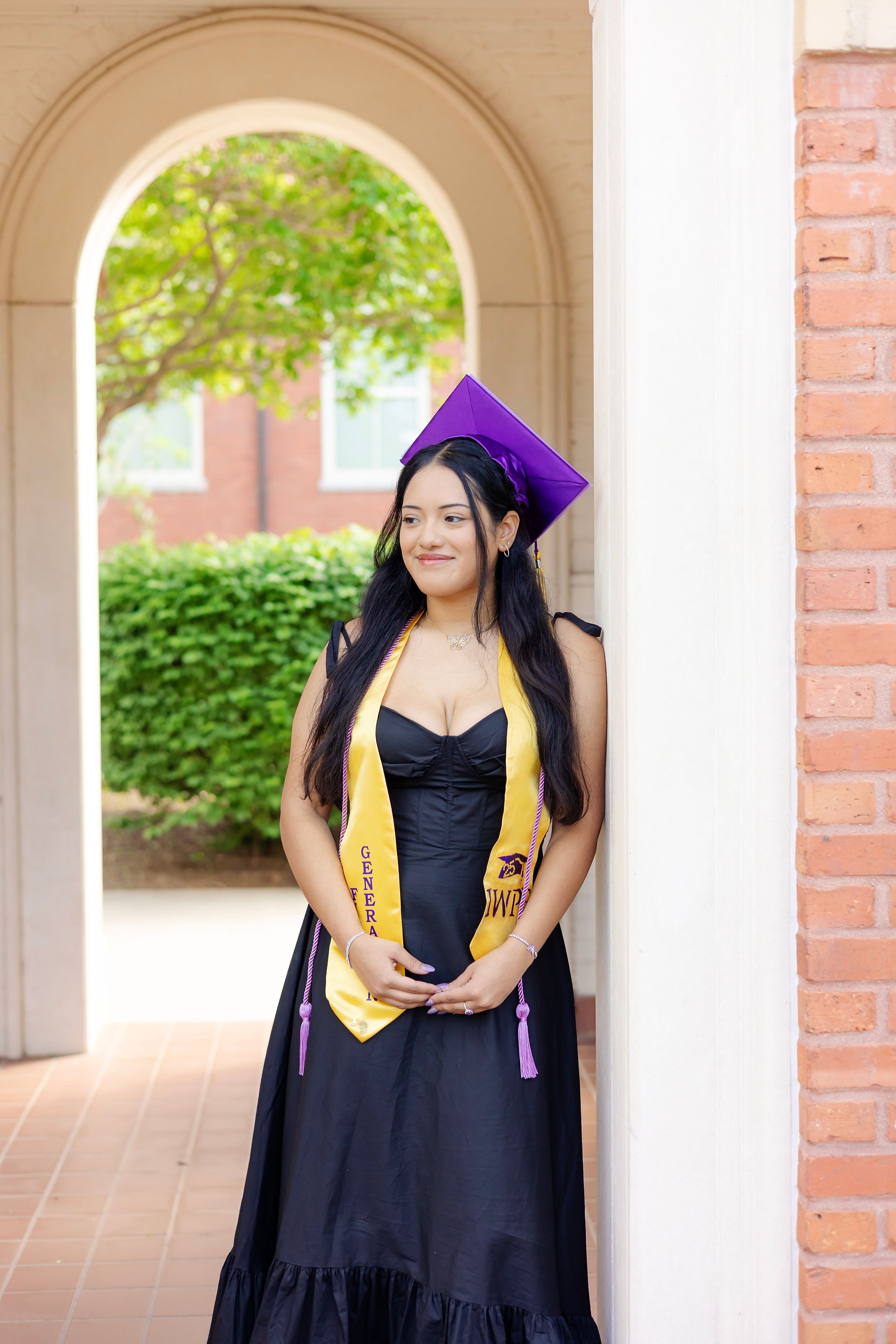 ECU graduate in black dress and cap posing under brick archway on East Carolina University campus.