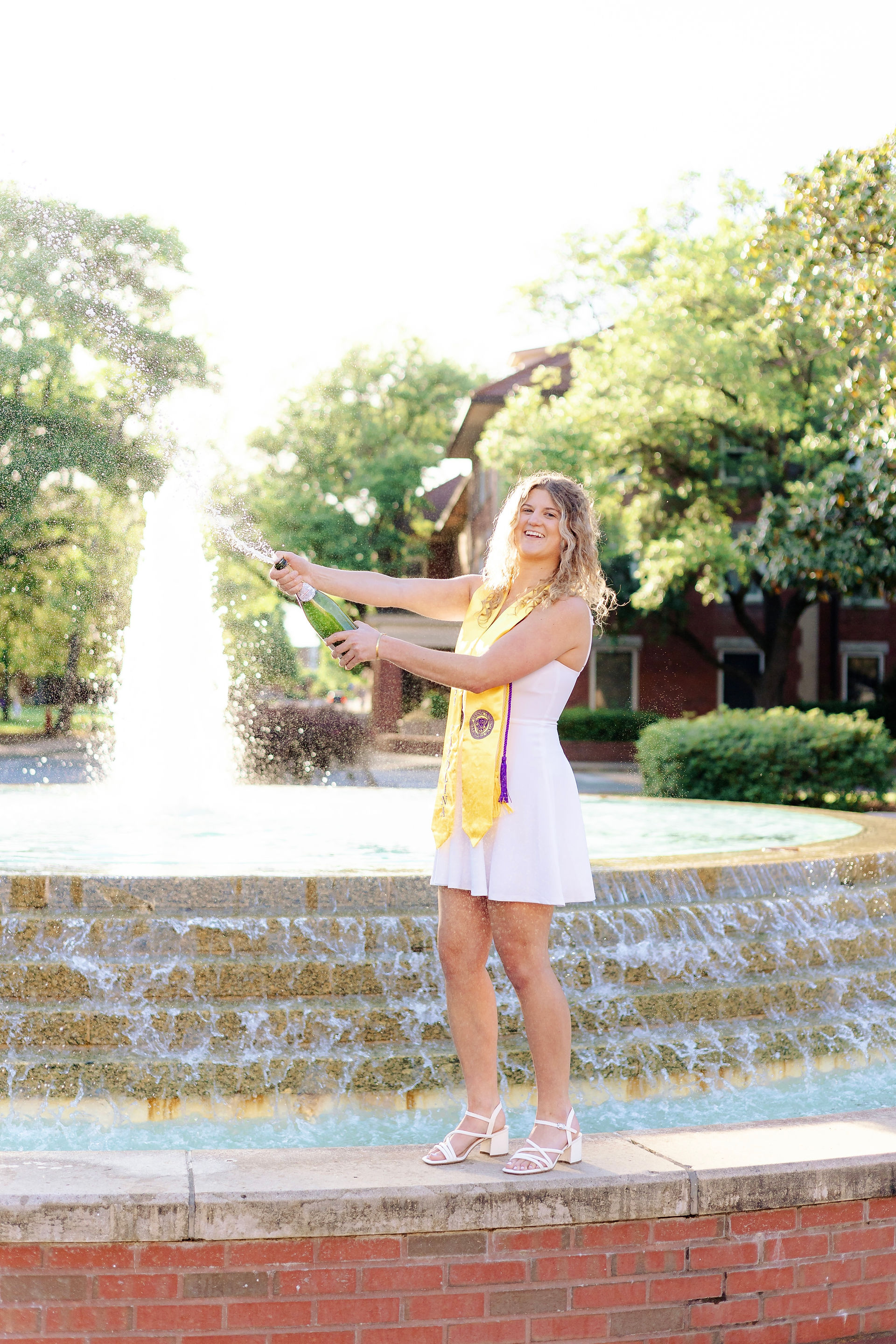 ECU senior popping champagne near campus fountain during graduation photo session.