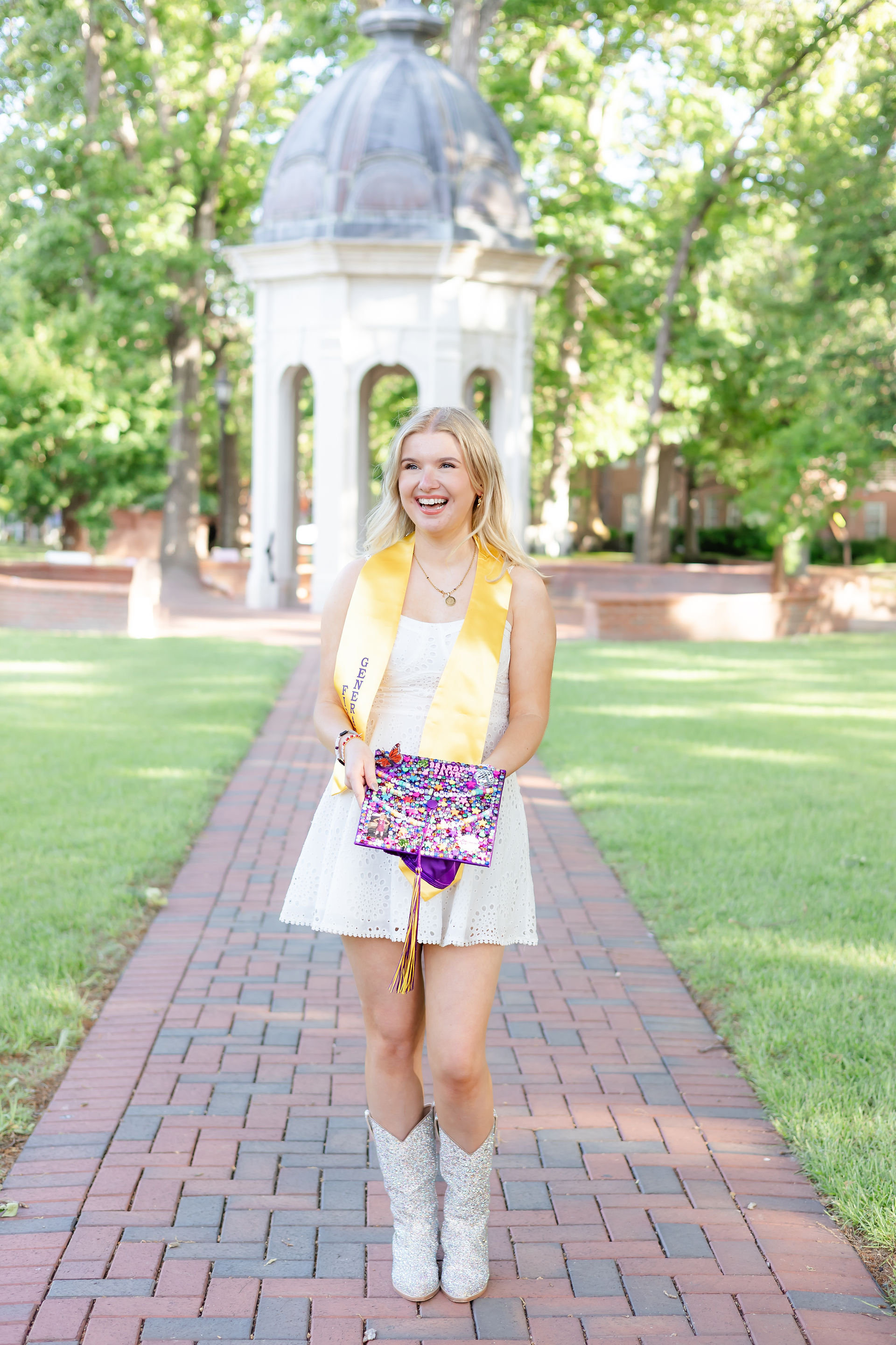 Female ECU senior walking on brick path toward the Cupola wearing white dress and decorated graduation cap.