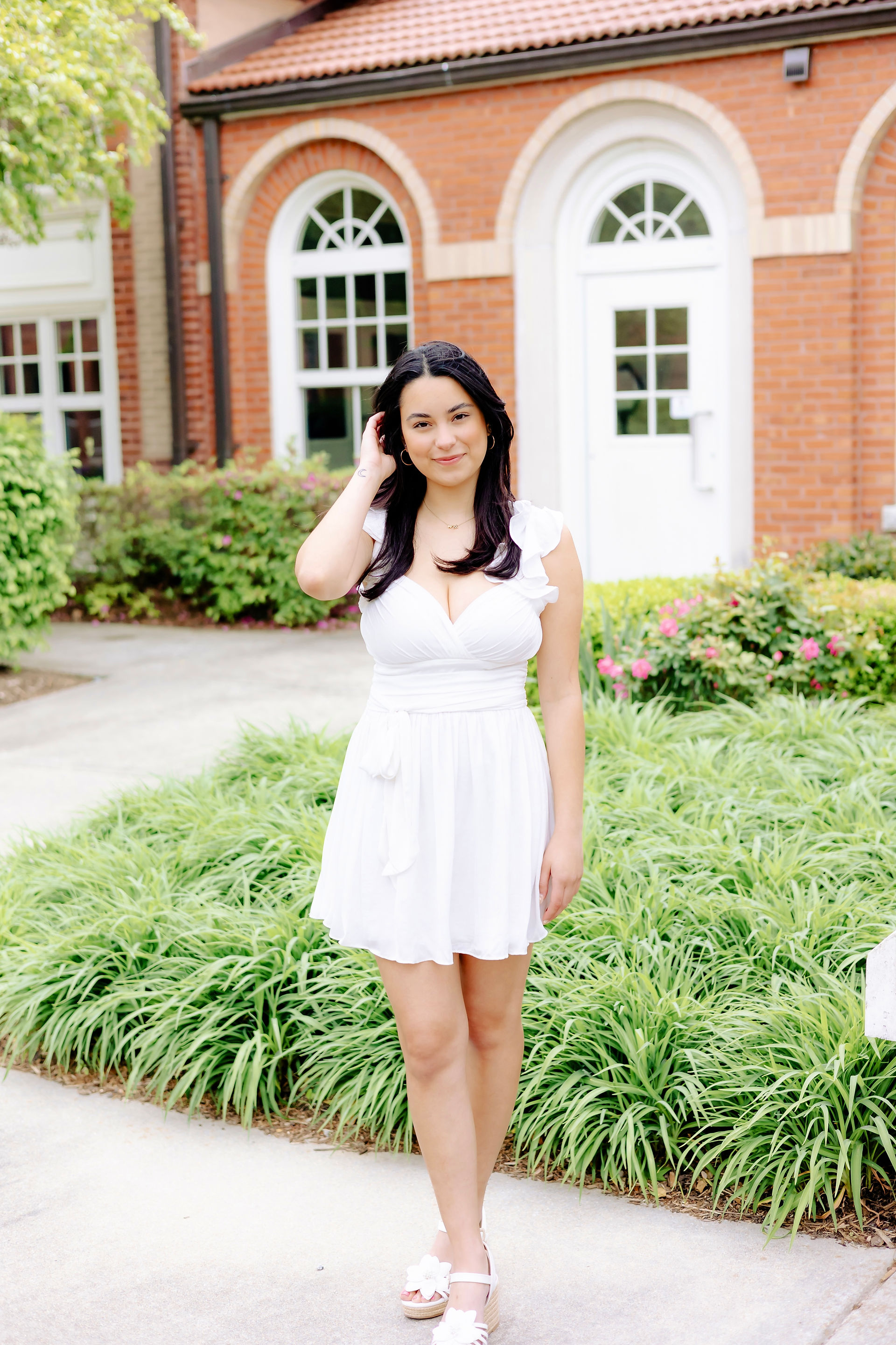 ECU senior in white dress posing near brick building and greenery at East Carolina University campus.
