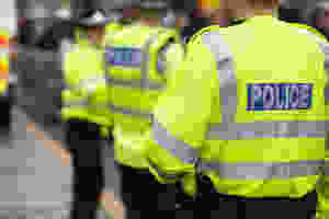 UK police offices one in focus with his back towards us. He is wearing a yellow high vis jacket with a blue box accross the shoulder blades which bears the wording Police.