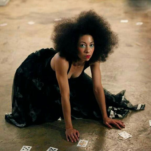 Stacy Lynn Smith, a mixed-race/Black dancing body with an Afro, looks out while kneeling in the middle of a circle of playing cards.