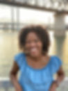 Milan, a young, Black woman smiles widely at the camera. She wears a jean-blue short-sleeved shirt. Her shoulder-length hair falls in a series of crinkles. Behind her is a bridge. The setting sun bathes the river in a warm amber glow.