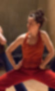 Donna, a white long limbed female dancer, is wearing an orange ribbed tank top, burnt orange loose pants and her hair is tied in a messy bun on top of her head. She is caught by camera looking pensively over her rolled forward right shoulder with the backs of her wrists on her waist, elbows and knees bent, and in a wide stance.