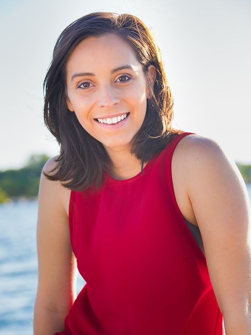 Paty gives a warm smile towards the camera. She has shoulder-length brown hair, olive skin, dark brown eyes. She is wearing a bright red, sleeve-less dress. The sun hits the right side of her face and upper torso as she sits against a blurred background of a lake and green grass.