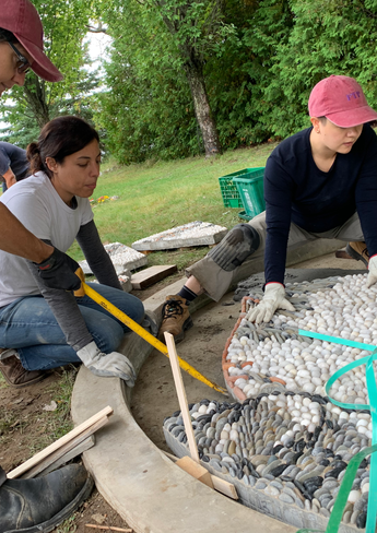 3 crew members gather around a large pebble mosaic they are installing. They use a crow bar and level to make adjustments as they work.