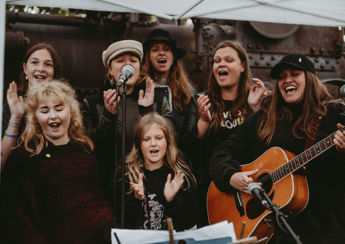 a group of singers mid-performance. One person plays an acoustic guitar.