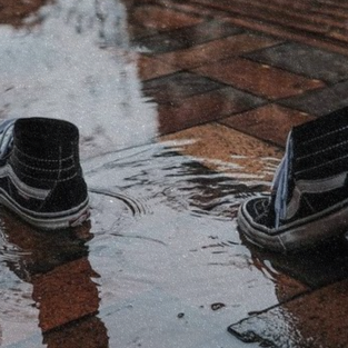 Two black sneakers stepping through a shallow puddle on a brick sidewalk.
