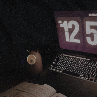 A dark, grainy photo of a laptop showing 12:55 beside a coffee mug at night.