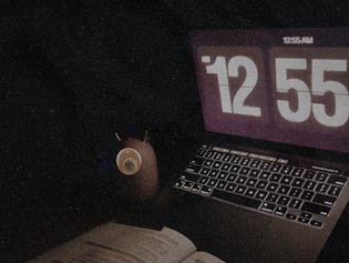 A dark, grainy photo of a laptop showing 12:55 beside a coffee mug at night.