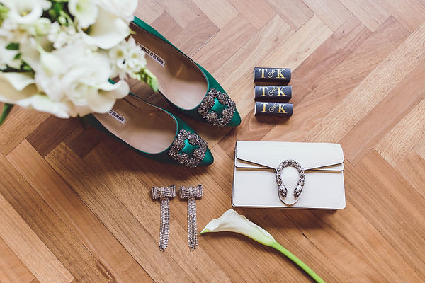 top-down image of the brides wedding day accessories featuring a green pair of shoes, bow silver earrings, a white clutch and 3 matchboxes that say, "T & K"