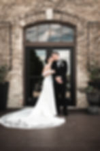 Bride and groom kiss in front of a rustic brick wall and arched window. She wears a white gown; he’s in a black tux. Romantic setting.