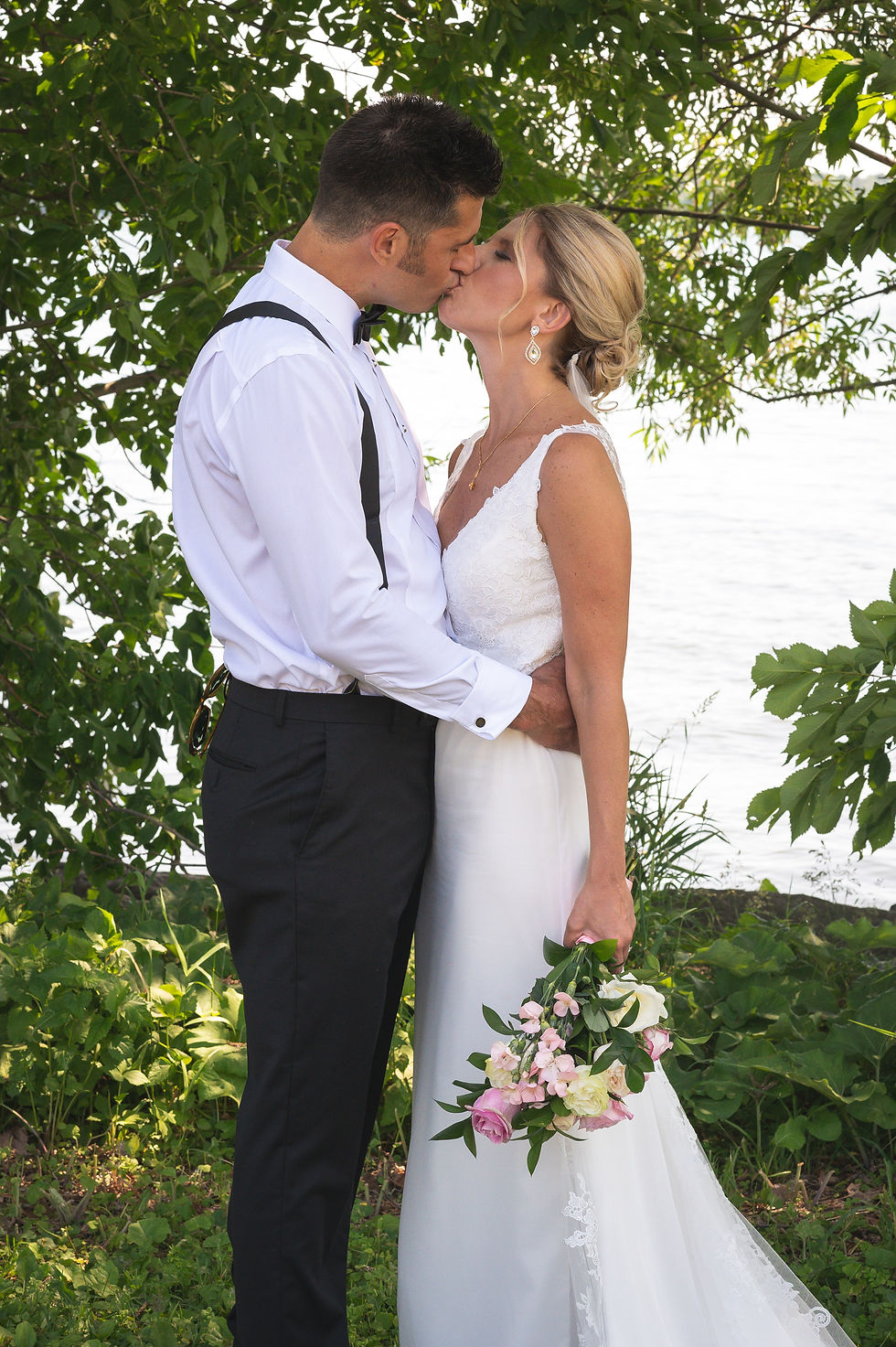 Groom kissing bride with his arms around here in front of lake and trees