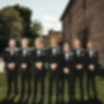 Seven men in black tuxedos pose smiling in a sunny outdoor setting. They stand on grass, with a brick building and trees in the background.