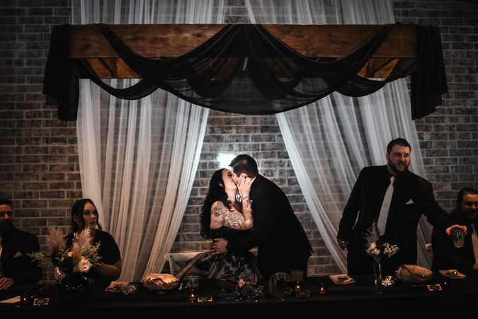 bride in black dress and groom in black suit kissing at head table during wedding reception at Whispering Pines Event Center in Poynette Wisconsin