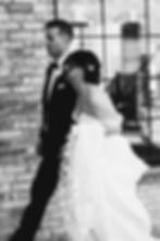 Bride and groom walk by a brick wall. The bride wears a floral gown, and both look joyful. Black and white photo with no visible text.
