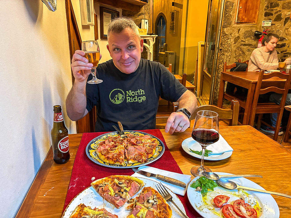 Man in black "North Ridge" shirt toasts with beer at a table with pizza and wine. Cozy restaurant setting with stone walls. Parque Natural da Serra da Estrela, Manteigas, Beira Alta, Portugal