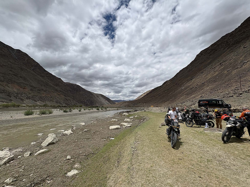 Motorcyclists gather beside a river in a rocky mountain valley under dramatic clouds, with an expedition van nearby. AvVida.co.uk - Life is an Adventure / La Vida es Una Aventura