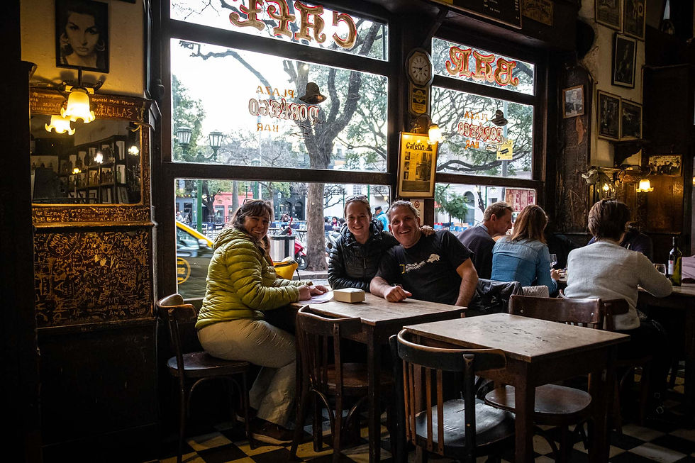 People smiling at a wooden table in a cozy café. Graffiti-covered walls, large windows revealing a street view, framed by warm lights and decor. AvVida.co.uk Adventure Motorcycle Travel South America Buenos Aires, Argentina