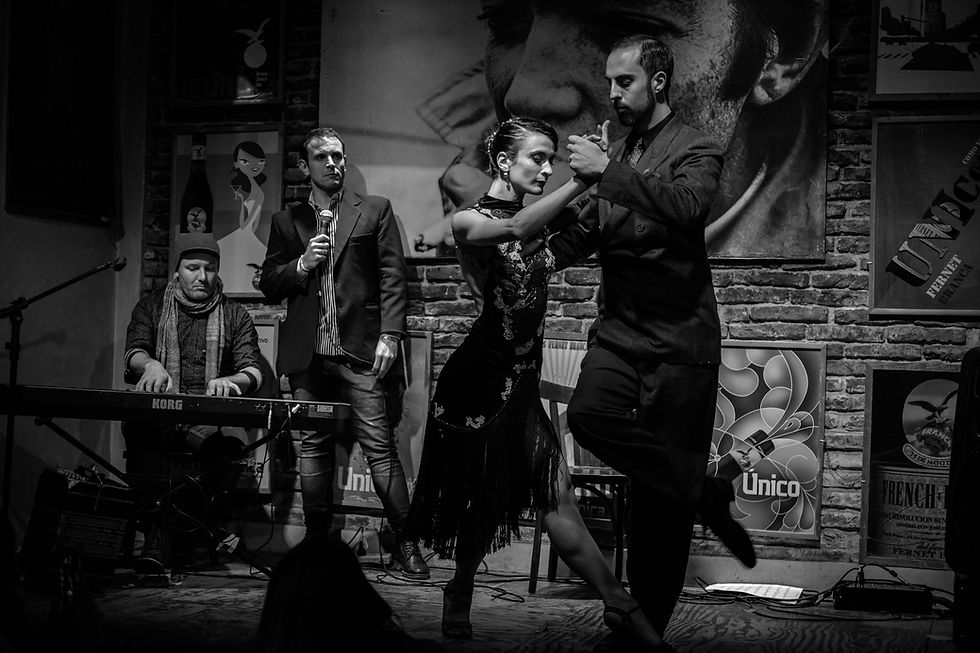 Dancers perform a tango in a dimly lit room with brick walls and posters. A man sings into a microphone, and another plays a keyboard. AvVida.co.uk Adventure Motorcycle Travel South America Buenos Aires, Argentina