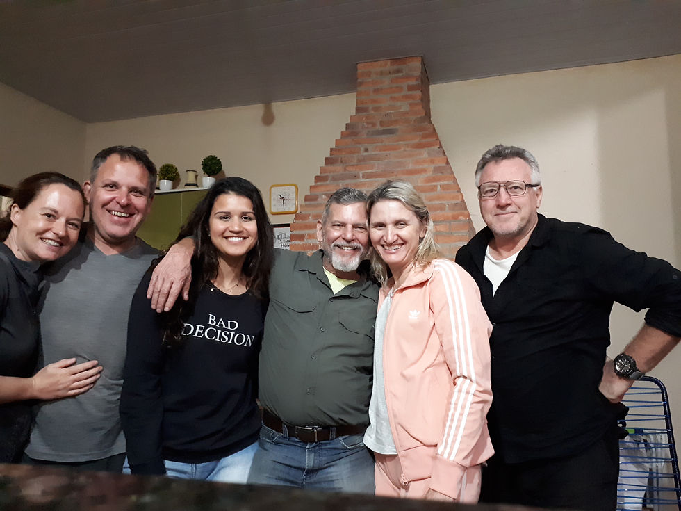 Six smiling adults stand close indoors. One wears a "Bad Decisions" shirt. Background has a brick chimney and plants. Casual, cheerful mood. AvVida.co.uk - Adventure Motorcycle Travel South America