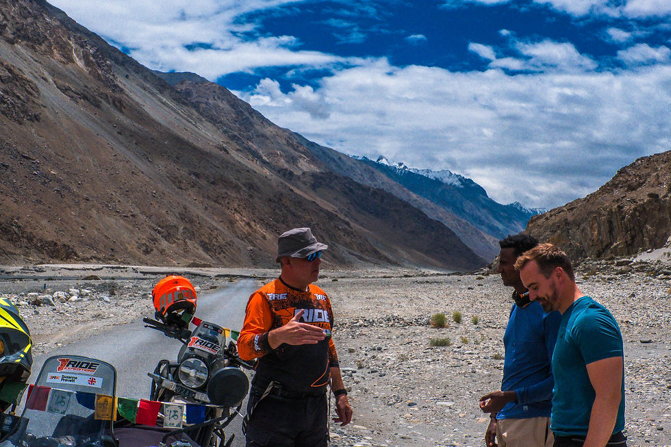 Three men chat beside a loaded motorcycle on a rocky mountain pass under a cloudy blue sky. AvVida.co.uk - Life is an Adventure / La Vida es Una Aventura