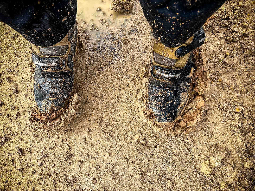Two muddy boots standing on wet, sandy ground. The boots are covered in dirt, creating a rugged and earthy mood. AvVida micro adventures – Life is an Adventure. Adventure Motorcycle Travel. Morocco. 