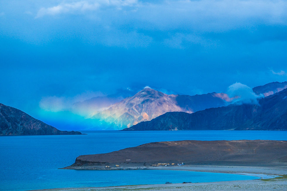 Blue lake and rugged mountains under cloudy sky, with a sandy shore and tiny lakeside buildings in a serene scene. AvVida.co.uk - Life is an Adventure / La Vida es Una Aventura